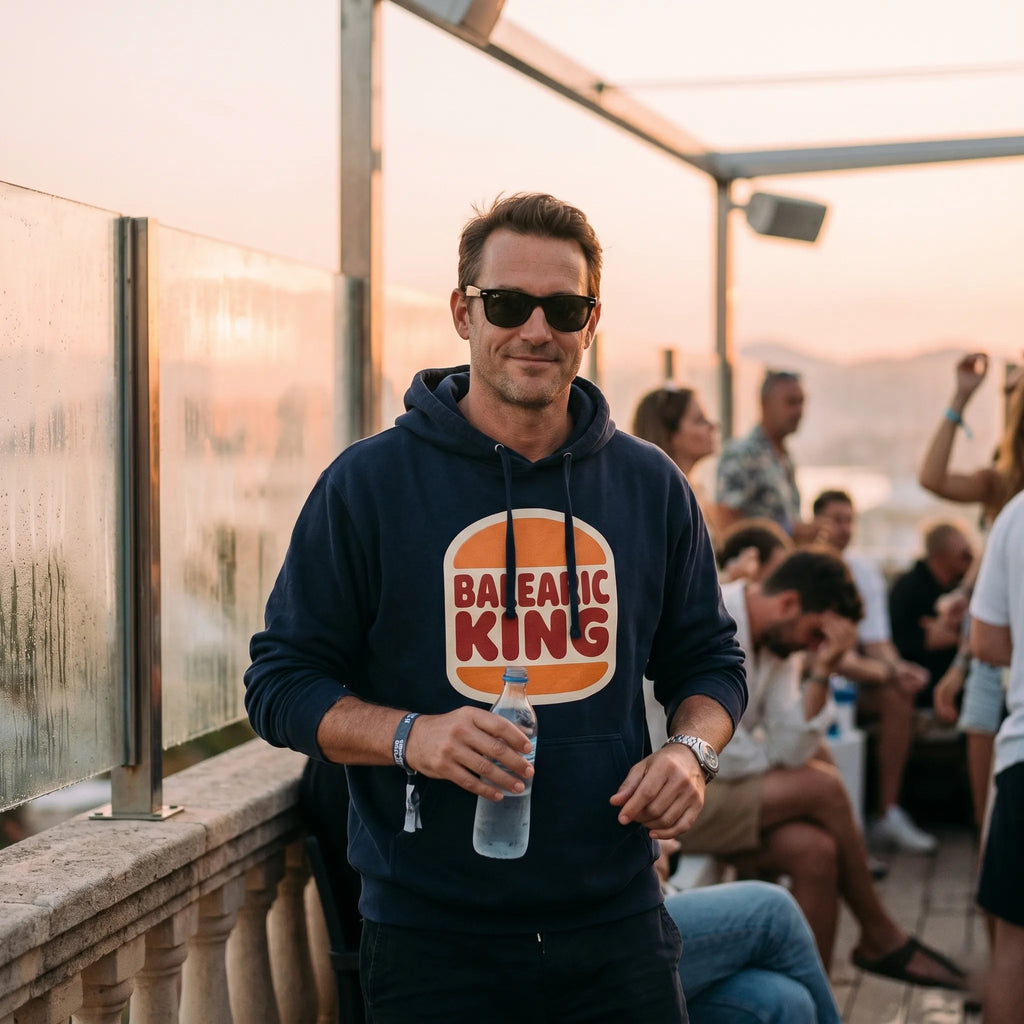 Man wearing a hoodie with 'Balearic King' design, holding a bottle, on a rooftop with people in the background.