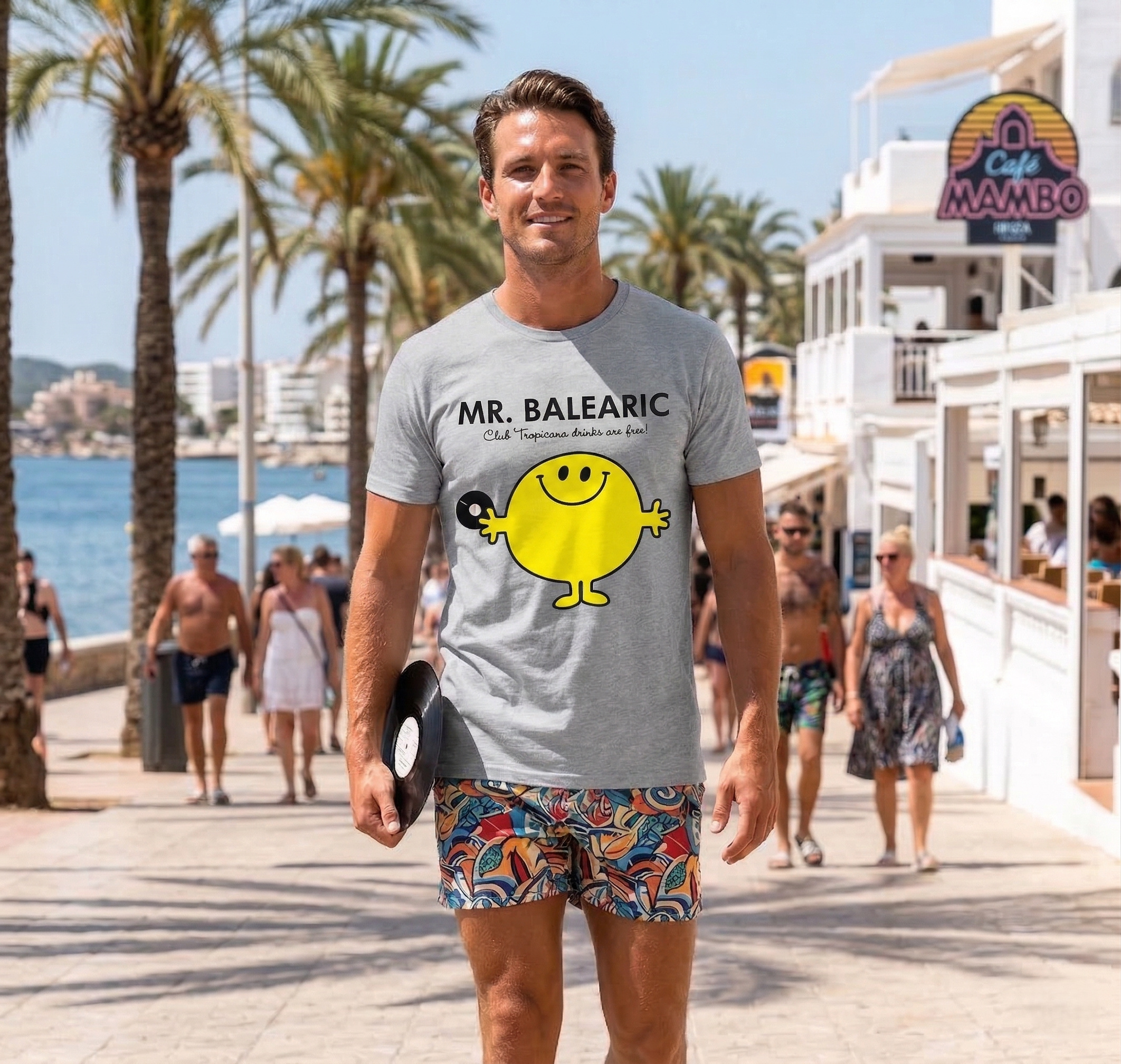 Balearic Blend T-shirt: Man in a grey Mr. Balearic graphic tee holding a vinyl record, walking along the sunny Ibiza waterfront near Café Mambo.